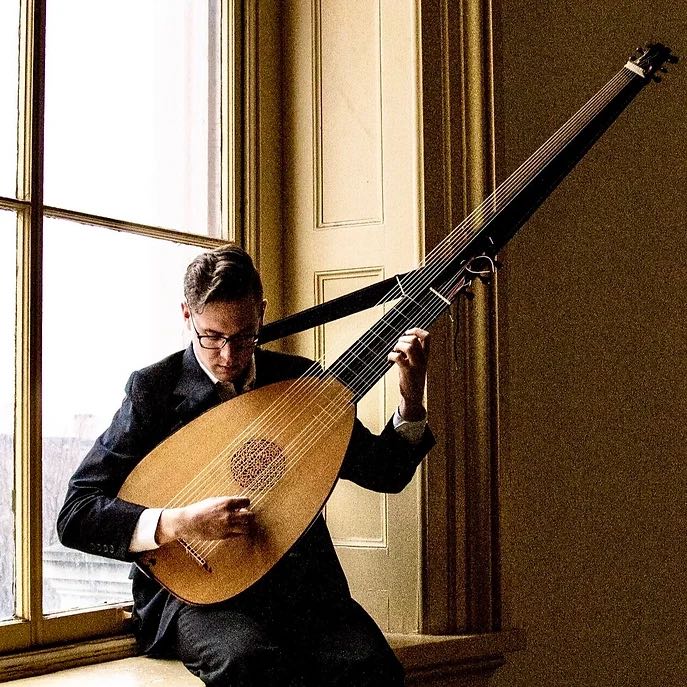 younger man sitting on a windowsill playing a large lute