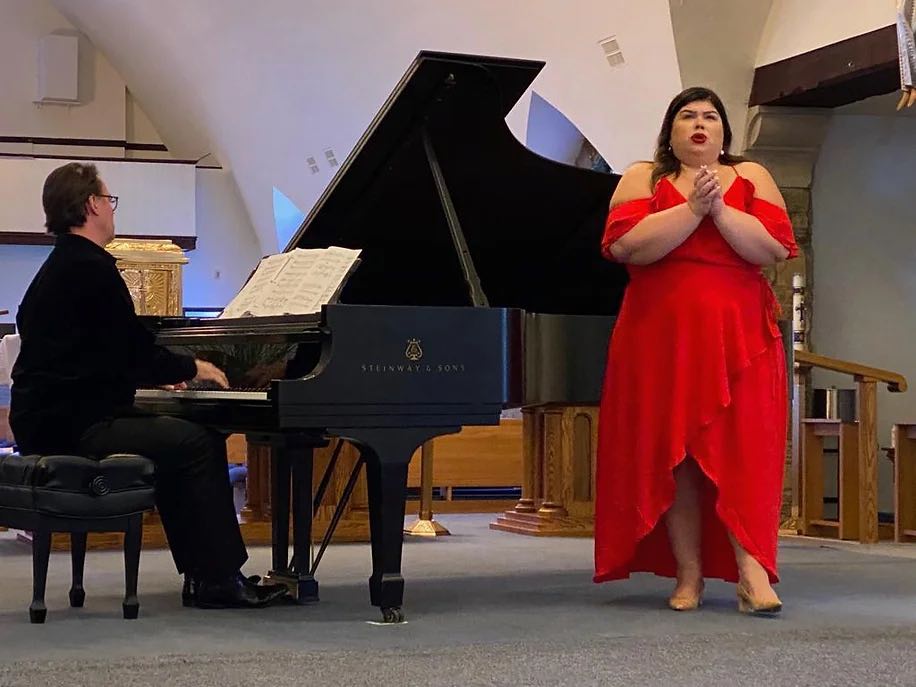 opera singer in a red dress with her hands together and mouth open next to a pianist playing