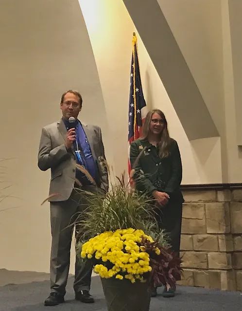 man speaking into microphone and woman holding her hands together next to him, both behind a large potted plant