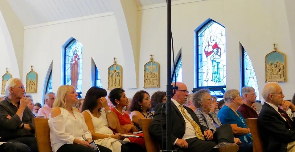 concert goes sitting in pews of a church with stained glass windows in the background