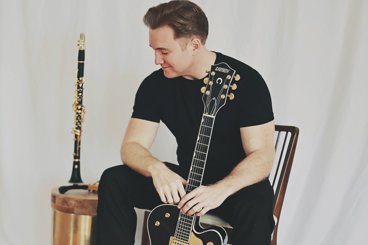 Dave Bennett sitting on a chair and posing with his guitar and clarinet