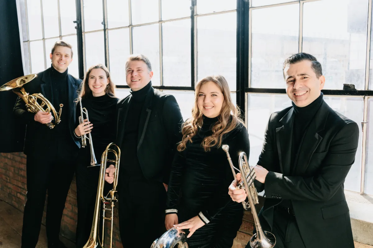 5 members of Axiom Brass holding there instruments wearing all black and standing in front of large windows