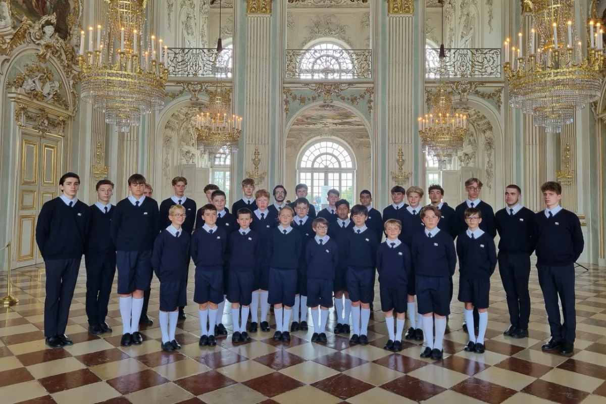 The French boys choir stands composedly in a massive, ornate palace hall beneath sparkling crystal chandeliers and gilded decorations.