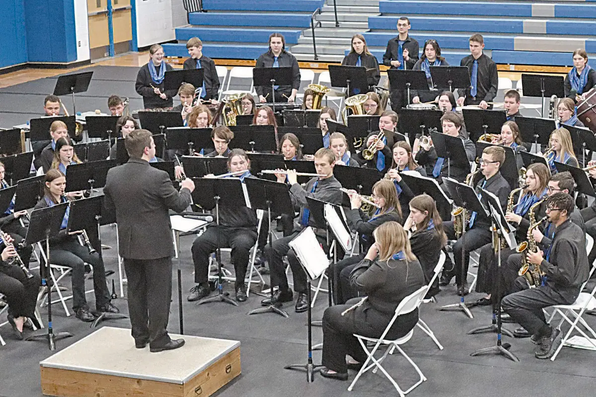 The conductor of the Cros-Lex Symphonic Band directs dozens of students dressed in black and blue uniforms, seated with instruments and music stands in a large gymnasium.