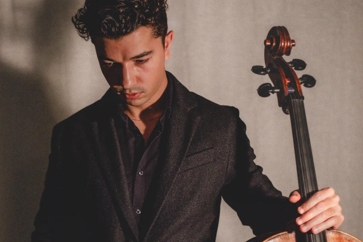 Cellist Gabriel Martins in a dark blazer holds his instrument and looks down with a serious, contemplative expression in dramatic lighting.