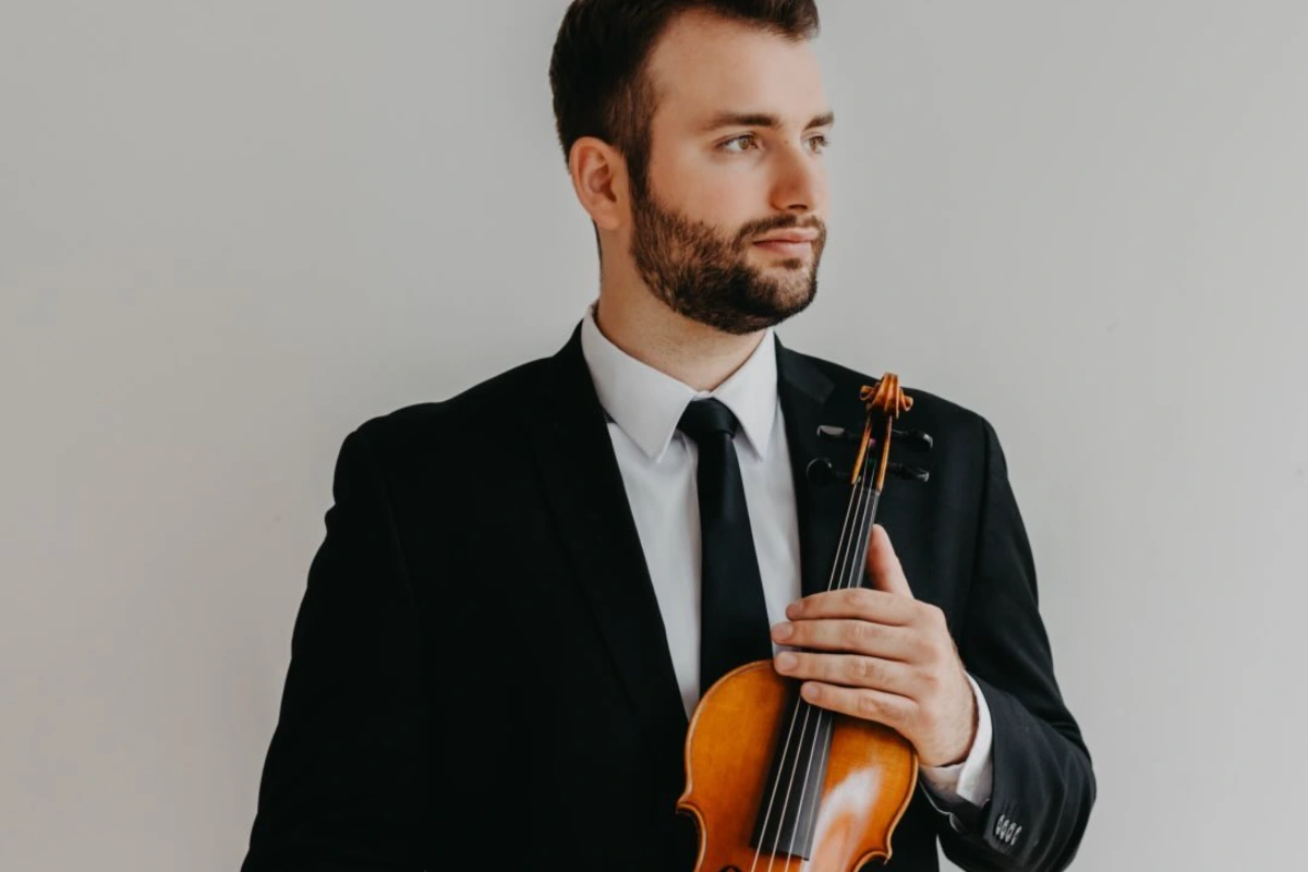 Konrad Kowal, wearing a black suit, white shirt, and black tie, holds a brown violin and looks thoughtfully to the side.