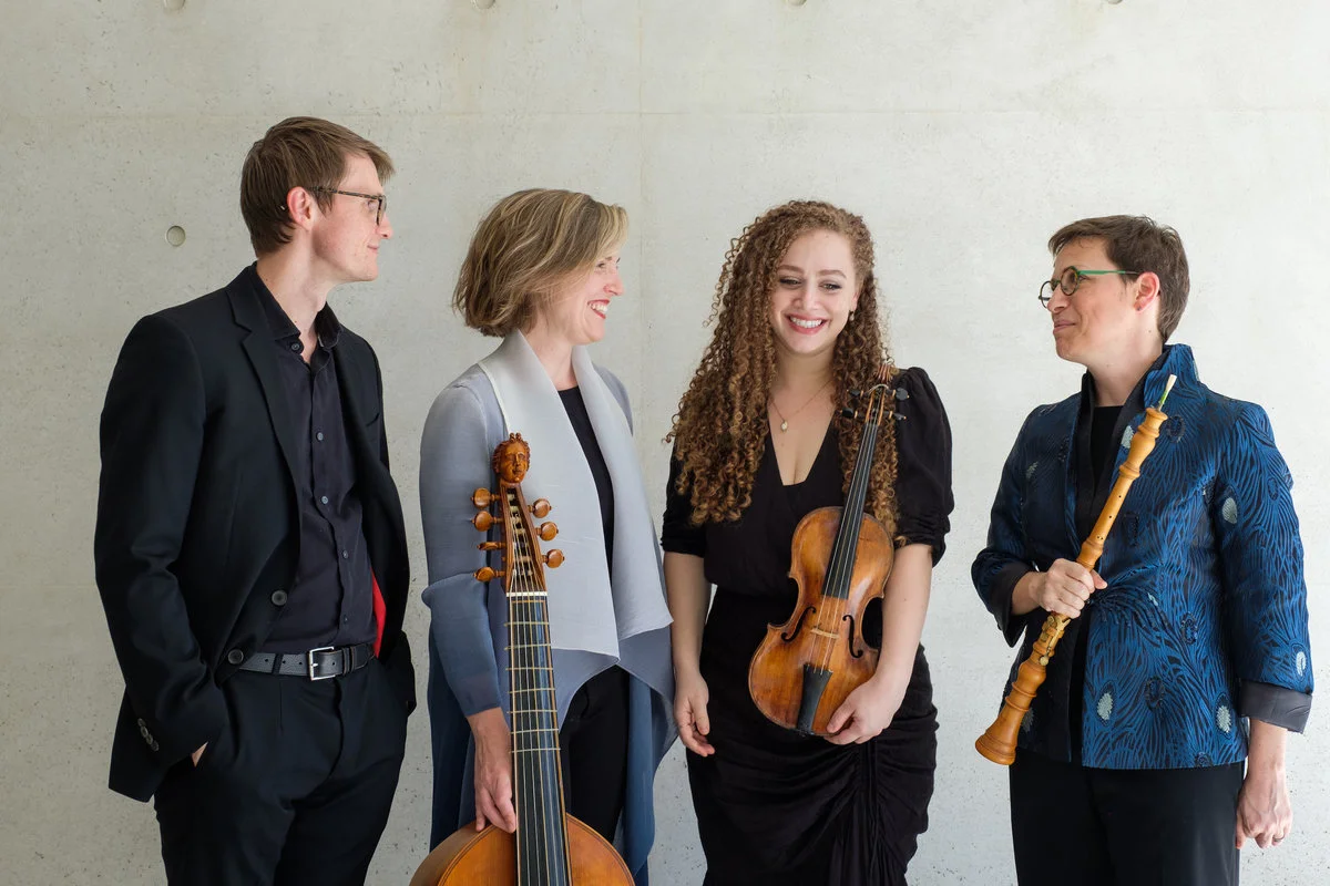 Four musicians, smiling and looking at each other, stand for a portrait holding a violin, a viola da gamba, and a baroque oboe.