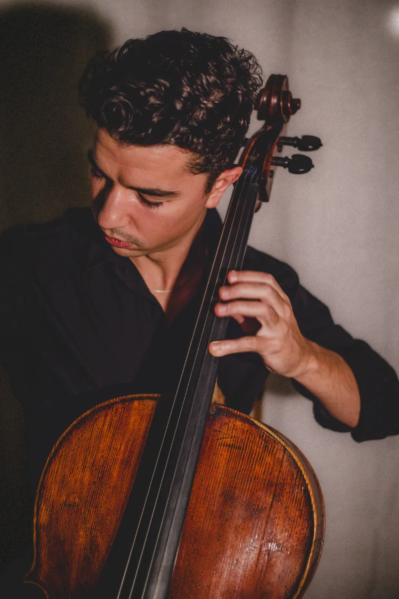 Celloist Gabriel Martins with dark, curly hair plays a wooden cello, concentrating intently on his performance in a dimly lit setting.