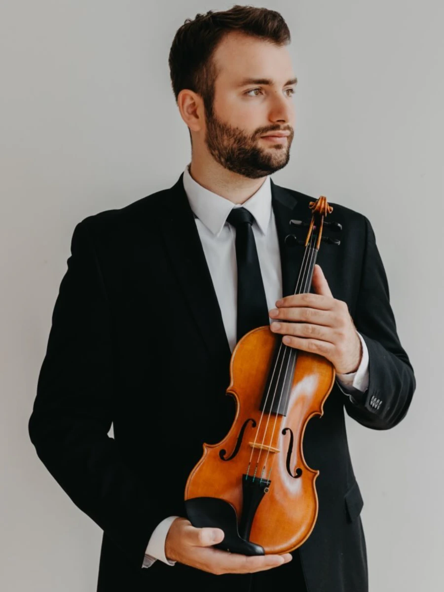 Violinist Konrad Kowal in a dark suit and tie holds his instrument and looks away with a focused expression.